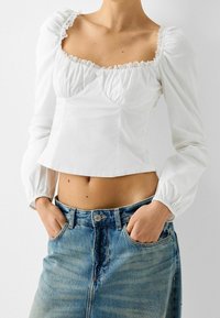 Blusa bianca corta con scollatura quadrata, bordo in pizzo e maniche a sbuffo, abbinata a jeans in denim blu a vita alta con una texture sbiadita.