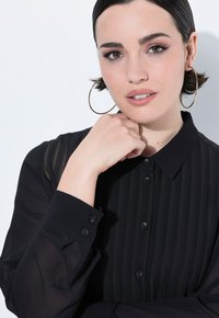 Zwarte, langmouwige button-up blouse met gestreepte textuur, kraag en doorzichtige stof bij de mouwen. Model draagt grote gouden hoepelringen.