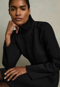 Schwarzes, tailliertes Kleid mit hohem, geripptem Kragen und Reißverschluss. Glatte Stoffqualität, lange Ärmel und sichtbare Nahtdetails an den Seiten.