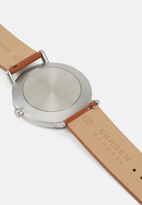 Silverfärgad klockkapsel i rostfritt stål med ett beige läderarmband. Lädret är präglat med "SKAGEN DANMARK" och har en slät yta.
