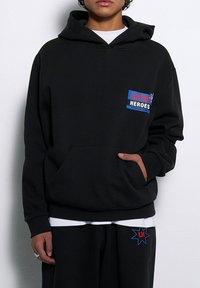 Czarna bluza z kapturem wykonana z miękkiego materiału, z przednią kieszenią, regulowanym kapturem oraz kolorową grafiką na klatce piersiowej z napisem "LOKALNI BOHATEROWIE".