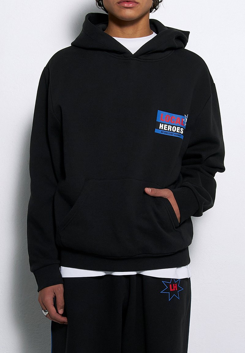 Czarna bluza z kapturem wykonana z miękkiego materiału, z przednią kieszenią, regulowanym kapturem oraz kolorową grafiką na klatce piersiowej z napisem "LOKALNI BOHATEROWIE".