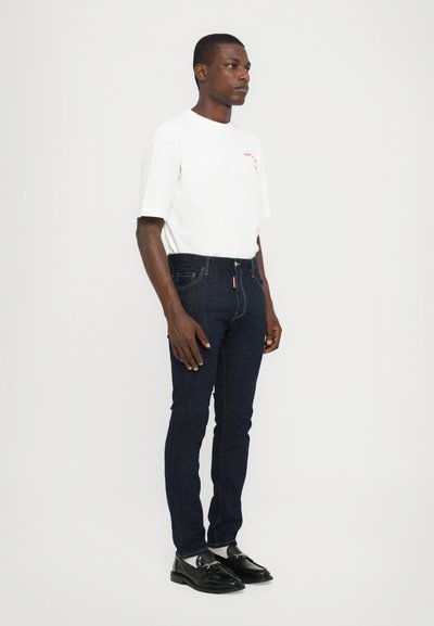 White crew-neck t-shirt with red graphic, paired with dark blue slim-fit jeans and black loafers. Model stands against a plain white background.