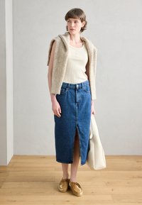 Gilet beige a maglia sopra una canotta a coste bianca, abbinato a una gonna di jeans blu con uno spacco frontale e scarpe marroni. Una borsa a spalla color crema è nei paraggi.