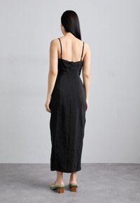 Robe maxi en lin noir avec bretelles réglables, corsage ajusté et silhouette droite. Présente une texture subtile et une fermeture à bouton à l'arrière.