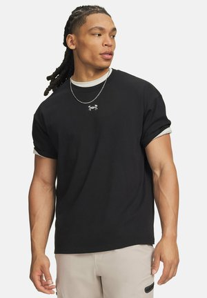 Jonge man met dreadlocks die een zwart Under Armour T-shirt draagt over een wit overhemd en beige broek, kijkt naar de zijkant.
