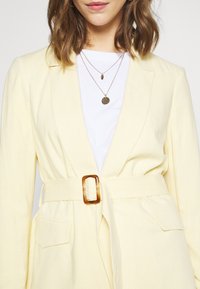 Yellow linen blazer with a belted waist, featuring a transparent buckle. White T-shirt underneath, layered with delicate gold necklaces.