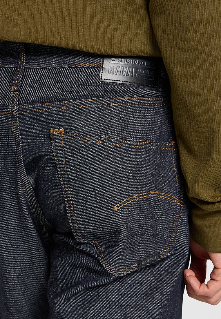 Dunkelblaue Denim-Jeans mit orangefarbenen Nähten, ausgestattet mit einem Lederpatch am Bund und einer Gesäßtasche mit einer geschwungenen Form.