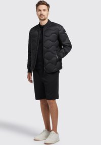 Black puffer jacket with wave pattern, zipper closure, paired with a black T-shirt and shorts. Model wearing white sneakers.