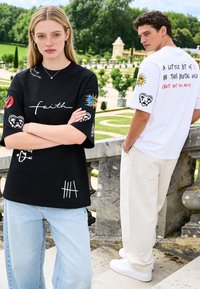 Jeune femme en t-shirt noir "faith" et jean, les bras croisés, se tient devant un jeune homme en t-shirt blanc à motif graphique et pantalon clair en extérieur.