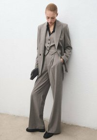 Traje gris de tres piezas con chaqueta a medida, chaleco con botones y pantalones de pierna ancha, acompañado de una camisa negra y un clutch.