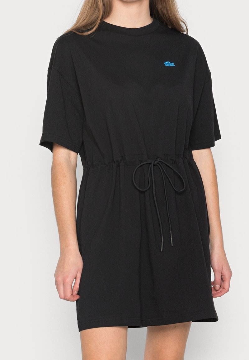 Femme portant une robe noire à manches courtes avec une taille à cordon et un petit logo bleu sur la poitrine sur un fond uni.