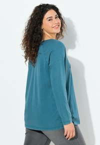 Tyrkysový top s dlouhým rukávem a volným střihem, kulatým výstřihem a šitými detaily. V kombinaci s tmavě šedými kalhotami v neformálním prostředí.