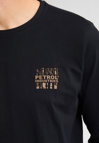 Schwarzes Langarm-T-Shirt aus Baumwolle mit einem goldenen Logo "PETROL INDUSTRIES" auf der linken Brustseite. Mit Rundhalsausschnitt.