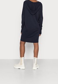Robe sweat à capuche surdimensionnée bleu marine avec manches longues, maille texturée, poignets côtelés et capuche au dos. Associée à des bottines crème à semelle épaisse.