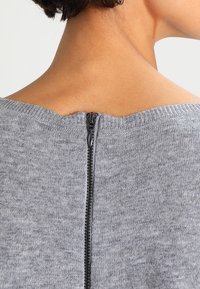 Grijs sweater met een ronde hals en een ritssluiting aan de achterkant met zwarte hardware. De stof heeft een zachte textuur met een gladde afwerking.