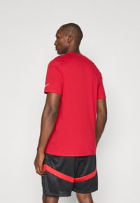Red cotton t-shirt with short sleeves and a round neckline, worn with black mesh shorts featuring red striped accents.