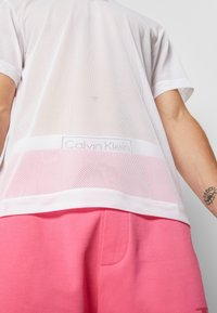 Bílý síťovaný tričko s krátkým rukávem se znakem na náprsní kapse. V kombinaci s růžovými šortkami, které mají jemnou texturu a ležérní styl.