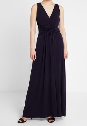 Mouwloze zwarte jurk tot op de grond met diepe V-hals en een knoopdetail in de taille, gedragen met zwarte open-toe hakken.