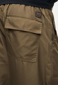 Κοντινή λήψη τσέπης καφέ παντελονιού cargo με καπάκι και ραμμένη ετικέτα που γράφει "TAION" στην πίσω περιοχή του γοφού.