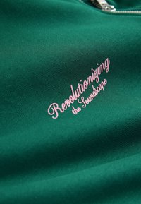 Grønt stof med lynlåslukning og rosa kursiv tekst, der lyder "Revolutionizing the Soundscape." Glat tekstur med minimal mønster.