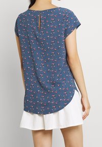 Blå blomsterkortärmad topp med nyckelhålsrygg, med små rosa och vita blommor på ett strukturerat tyg. Rundad fåll.