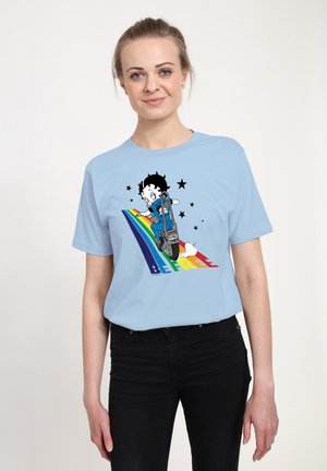 Lyseblå t-shirt med en tegneseriefigur med sort hår, der kører på en scooter på en regnbue, omgivet af stjerner og teksten "VÆR FRIE."