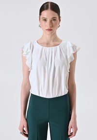 Donna che indossa una blusa bianca con rouches a maniche corte e pantaloni verde scuro a vita alta, in piedi davanti a uno sfondo chiaro e uniforme.