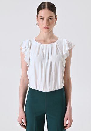 Woman wearing a white ruffled short sleeve blouse and dark green high-waisted pants, standing against a plain light background.