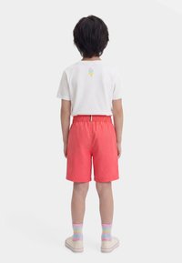 Παιδί που στέκεται με την πλάτη, φορώντας άσπρο μπλουζάκι με γραφικό παγωτού, κοραλί σορτς, παστέλ ριγέ κάλτσες και λευκά αθλητικά παπούτσια.