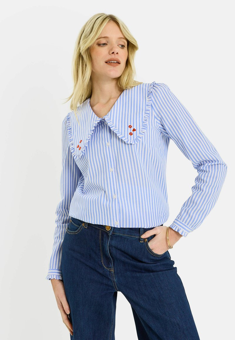 Femme blonde portant un chemisier à rayures bleues et blanches avec un grand col à volants et une petite broderie de fleurs rouges, associé à un jean foncé.