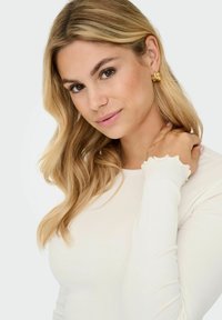 Witte geribde long-sleeve top met geschulpte manchetten, gestyled met losse golvende blonde haren en gouden creolen.