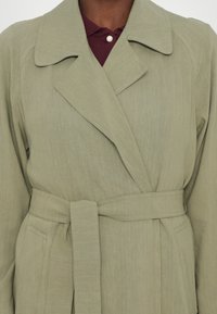 Cappotto trench belted verde oliva con colletto a punte, texture liscia e dettagli di cucitura orizzontale sulle maniche.