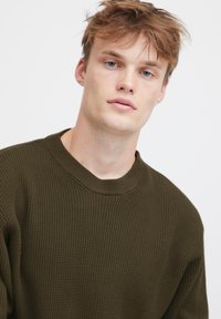 Oliven grønn teksturert genser med ribbestrikket hals og løs passform. Stoffet har et vaffelmønster som gir visuell interesse og dybde.