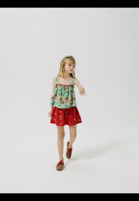 Ung pige iført en blomsterprintet ærmeløs top, rød nederdel med mønstre, pandebånd og brune sko, der går foran en ensartet hvid baggrund.