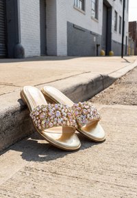 Złote sandały typu slip-on z odkrytymi palcami i dużymi, opalizującymi ozdobami z kamieni, spoczywające na nasłonecznionym krawężniku ulicy w pobliżu szarego budynku.