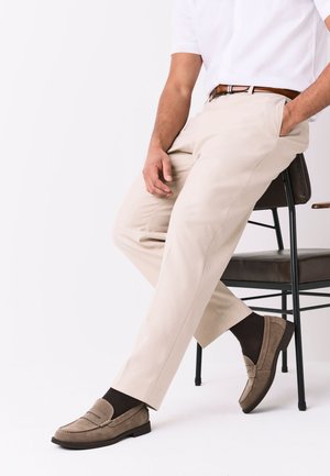 Mann sitzt auf einem schwarzen Stuhl, trägt beige Hose, braunen Gürtel, dunkle Socken und hellbraune Wildleder-Loafer, vor einem schlichten weißen Hintergrund.