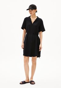 Schwarzes Button-Up-Kleid mit kurzen Ärmeln, taillierter Taille, aus strukturiertem Stoff. Kombiniert mit dunklen Rutsch-Sandalen und einer passenden Kappe.