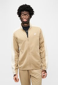 Μπεζ ζακέτα με φερμουάρ με εκρού λεπτομέρειες, κατασκευασμένη από λείο ύφασμα. Διαθέτει ψηλό γιακά, πλαϊνές τσέπες και μικρό λογότυπο Nike στο στήθος.