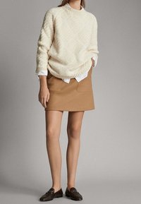 Pull en tricot crème avec un motif en losanges texturé, porté sur une chemise blanche à col. Mini-jupe marron avec deux poches avant, assortie à des mocassins foncés.