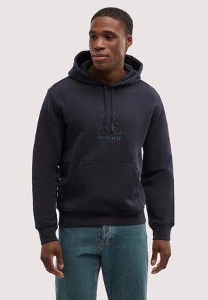 Mężczyzna ubrany w granatową bluzę z kapturem, kieszenią z przodu i logo, w połączeniu z niebieskimi dżinsami, patrzący lekko na bok.