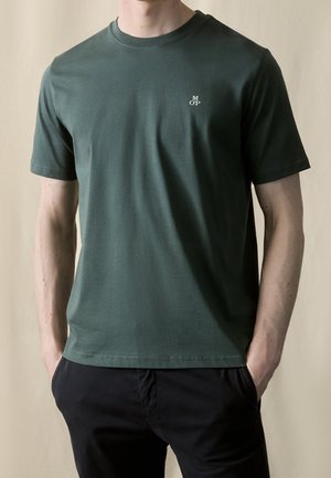 Homme portant un T-shirt à manches courtes vert foncé avec un petit logo blanc "MOP" sur la poitrine, les mains dans les poches de son pantalon noir.