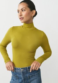 Olive-green, prążkowany golf z długimi rękawami, w zestawieniu z jasnoniebieskimi dżinsami o wysokim stanie, które mają przednie kieszenie i zapięcie na guzik.