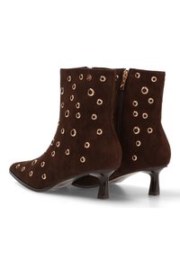 Botas de tobillo de ante marrón con puntera afilada, que presentan adornos de ojetes dorados circulares y una cremallera lateral, montadas sobre un tacón bajo y brillante.