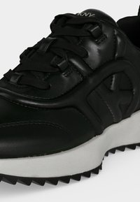Gros plan d'une basket en cuir noir avec des lacets noirs, logo embossé sur le côté, et une semelle blanche texturée avec une bande de roulement noire.
