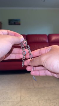 Μια μεταλλική ασημένια αλυσίδα με ωοειδή κρίκους και λεία υφή. Στις άκρες έχει μικρές πόρπες για κλείσιμο. Το φόντο περιλαμβάνει έναν κόκκινο καναπέ.