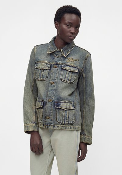 Άτομο που φορά ξεθωριασμένο μπλε τζιν μπουφάν με τέσσερις μπροστινές τσέπες και ανοιχτόχρωμο παντελόνι, στέκεται μπροστά σε απλό λευκό φόντο.