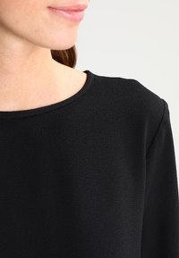 Schwarzes, strukturiertes Stoffoberteil mit rundem Ausschnitt und langen Ärmeln, das ein schlichtes Design und eine glatte Oberfläche aufweist.