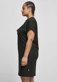 Robe en coton noir avec des manches roulées courtes, coupe droite, longueur genou, comportant une petite étiquette de marque verte sur le côté. Design simple.