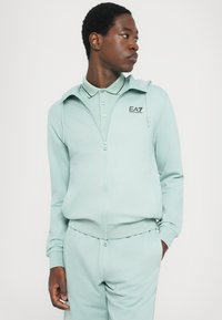 Lys myntegrønn zip-uphettegenser med matchende polo-krage. Har en logo på brystet, slim fit og myk stofftekstur.